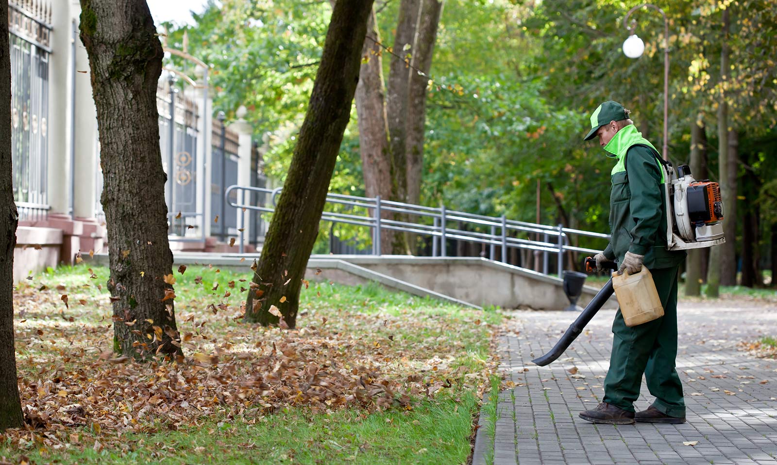 Gärtner in Arbeitsuniform entfernt mit einem Laubbläser Herbstlaub von einem Gehweg in einem städtischen Park.